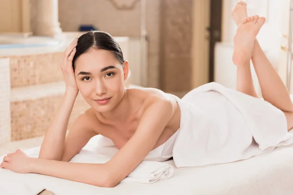 Hermosa mujer asiática en toalla acostado en mesa de masaje y mirando a la cámara - foto de stock