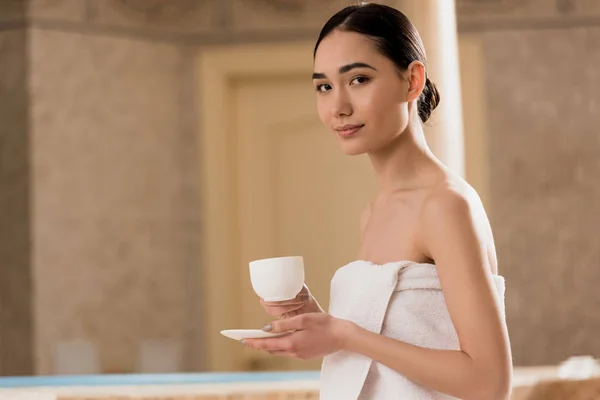 Hermosa mujer asiática en toalla sentado con taza de café y mirando a la cámara - foto de stock