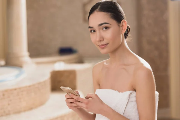 Attraktive asiatische Frau in Handtuch mit Smartphone und Blick auf die Kamera — Stockfoto