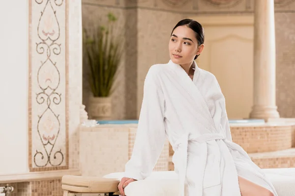 Hermosa mujer asiática en albornoz sentado y mirando hacia el spa - foto de stock