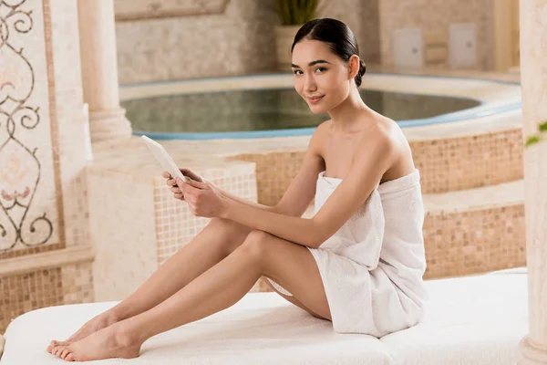 Atractiva mujer asiática en toalla usando tableta digital y mirando la cámara - foto de stock