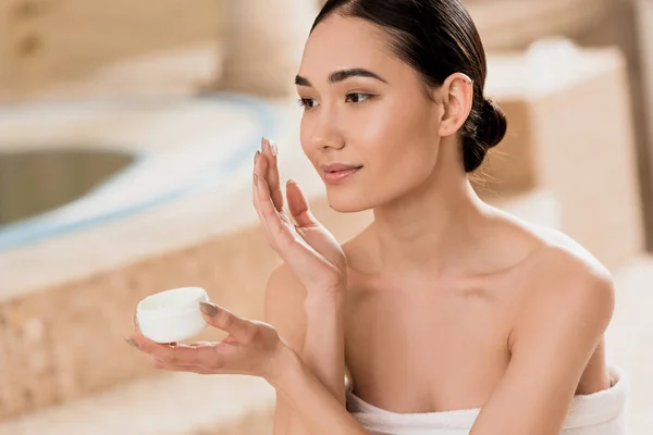 Attractive asian woman in towel applying cosmetic cream at spa — Stock Photo