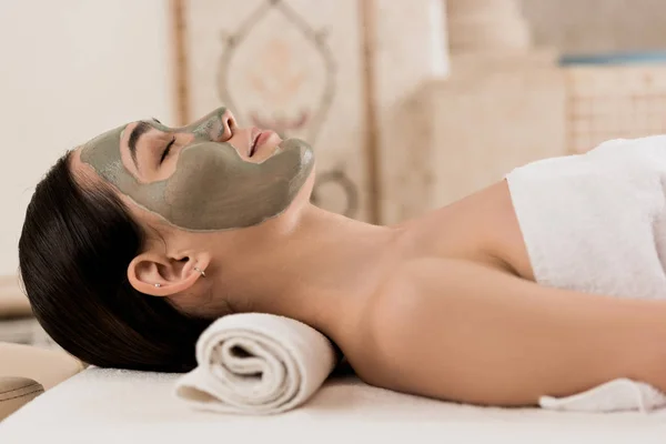 Vista laterale di donna attraente sdraiata con maschera di argilla sul viso al centro benessere — Foto stock