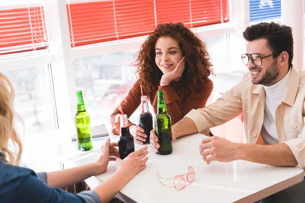 Улыбающиеся друзья, держащие стеклянные бутылки с содовой и пивом и разговаривающие — стоковое фото
