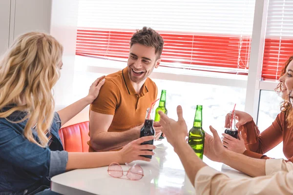 Smiling and beautiful friends holding glass bottles and talking at cafe — Stock Photo