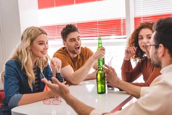 Selective focus of smiling friends holding glass bottles with drink and talking at cafe — Stock Photo