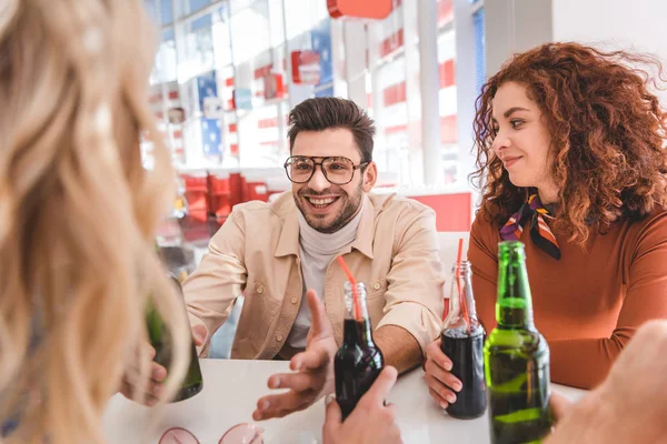 Selective focus of handsome and beautiful friends holding glass bottles with drink and talking at cafe — Stock Photo