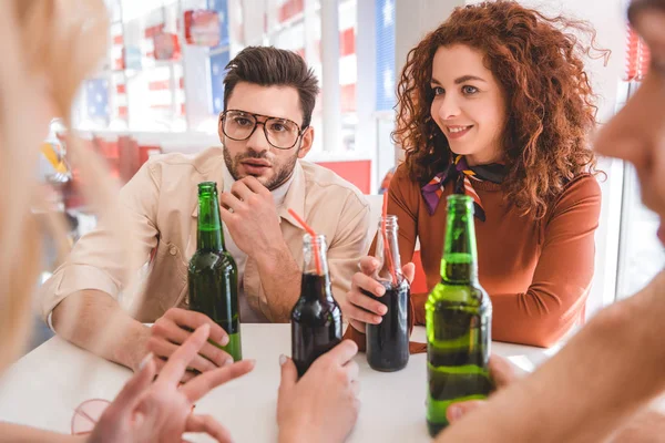 Selective focus of handsome and attractive friends holding glass bottles with drink and talking at cafe — Stock Photo