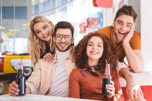 Guapos y hermosos amigos sosteniendo botellas de vidrio con bebida y sonriendo - foto de stock