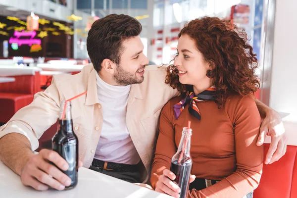 Casal sorrindo, segurando garrafas de vidro com refrigerante e olhando um para o outro — Fotografia de Stock