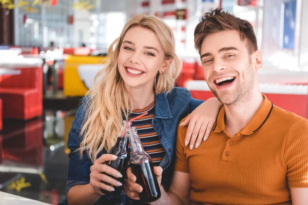 Hermosa pareja sonriendo, mirando a la cámara y animando con botellas de vidrio en la cafetería - foto de stock