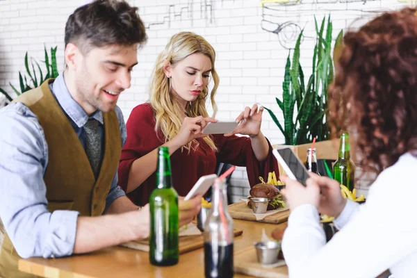 Selective focus of friends taking photo of burger and using smartphones at cafe — Stock Photo