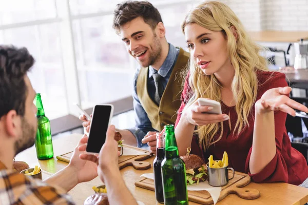 Enfoque selectivo de amigos hermosos y guapos hablando y usando teléfonos inteligentes en la cafetería - foto de stock