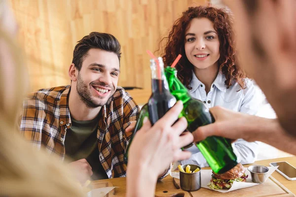 Selective focus of smiling and beautiful friends cheering with glass bottles at cafe — Stock Photo