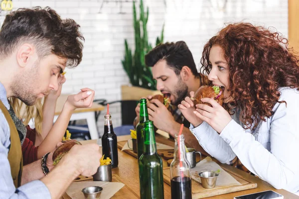 Enfoque selectivo de guapos y hermosos amigos comiendo sabrosas hamburguesas en la cafetería - foto de stock