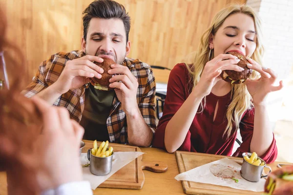 Selective focus of handsome and attractive friends eating delicious burgers at cafe — Stock Photo