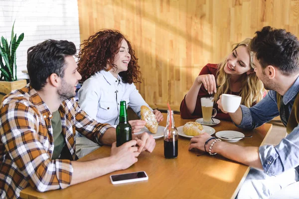 Guapos y hermosos amigos hablando, sonriendo y comiendo en la cafetería - foto de stock