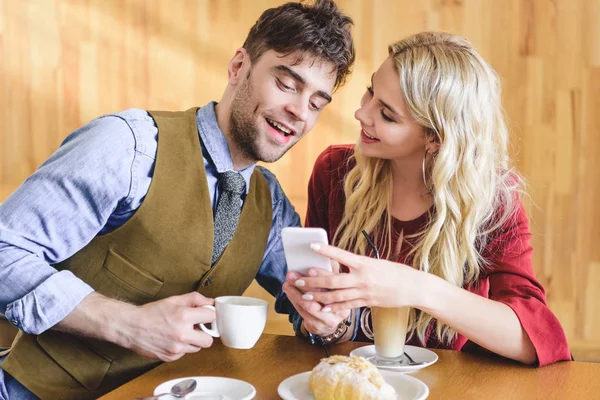 Hermosa pareja sosteniendo tazas de café y el uso de teléfono inteligente en la cafetería - foto de stock