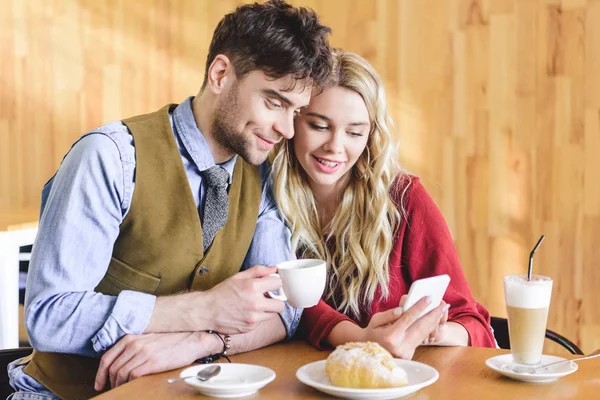 Bell'uomo che tiene in mano la tazza di caffè e la donna usando lo smartphone al caffè — Foto stock
