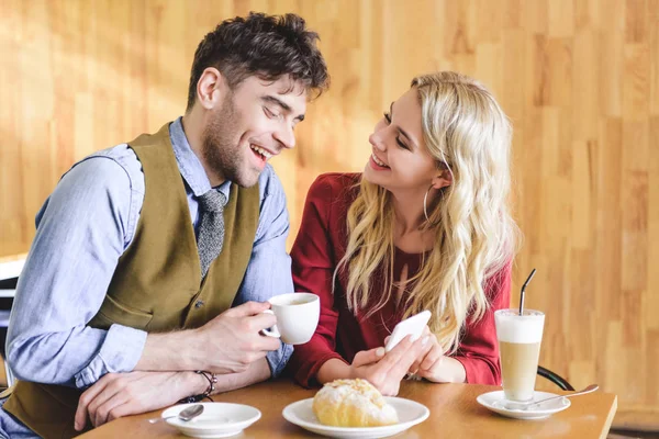 Bell'uomo con in mano una tazza di caffè e una bella donna che usa lo smartphone al caffè — Foto stock