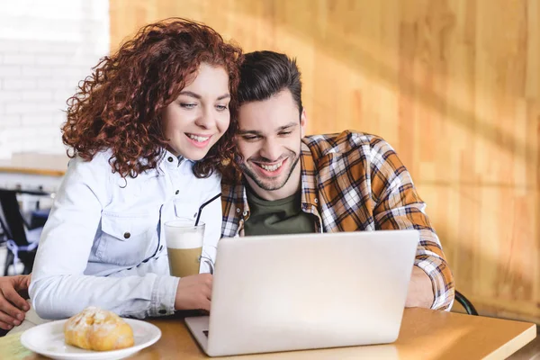 Beau et souriant couple étreignant et en utilisant un ordinateur portable au café — Photo de stock