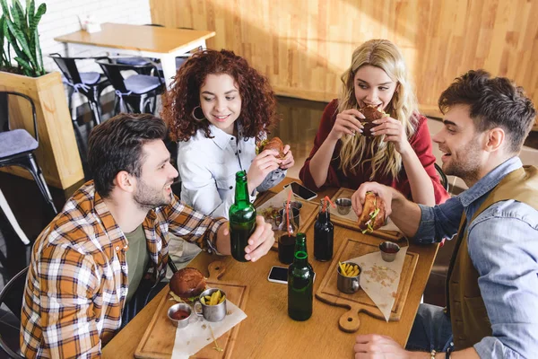 Guapos y hermosos amigos hablando, sonriendo y comiendo hamburguesas en la cafetería - foto de stock