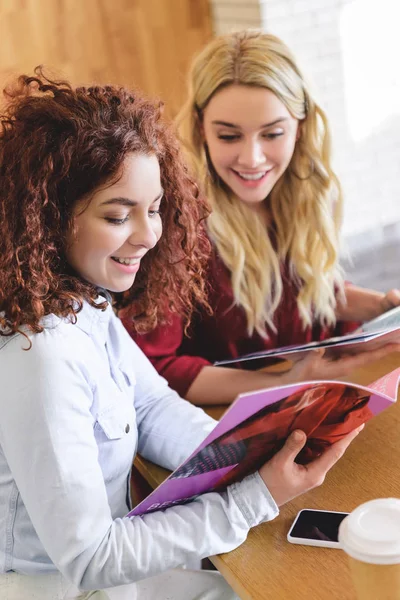 Selective focus of attractive and smiling women reading magazines and talking at cafe — Stock Photo