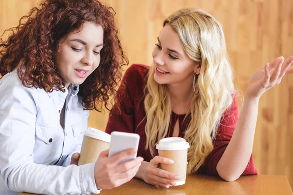 Mujeres atractivas y sonrientes usando teléfono inteligente y hablando en la cafetería - foto de stock