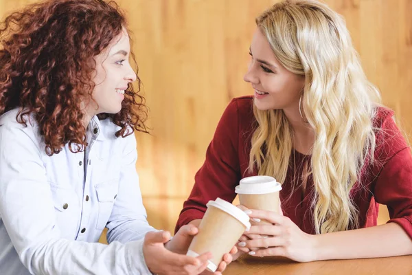 Mujeres hermosas y sonrientes sosteniendo vasos de papel y hablando en la cafetería - foto de stock