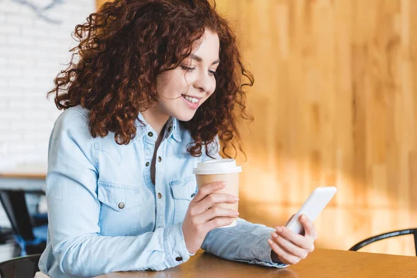 Красива і усміхнена жінка тримає паперову чашку і використовує смартфон — стокове фото