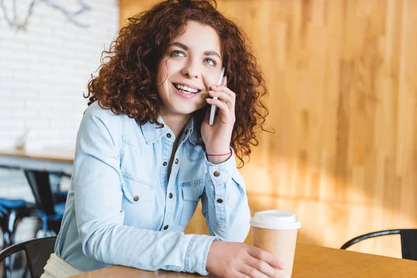 Hermosa y sonriente mujer sosteniendo taza de papel y hablando en el teléfono inteligente - foto de stock