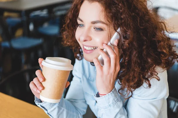 Привлекательная и улыбающаяся женщина держит бумажную чашку и разговаривает на смартфоне — стоковое фото