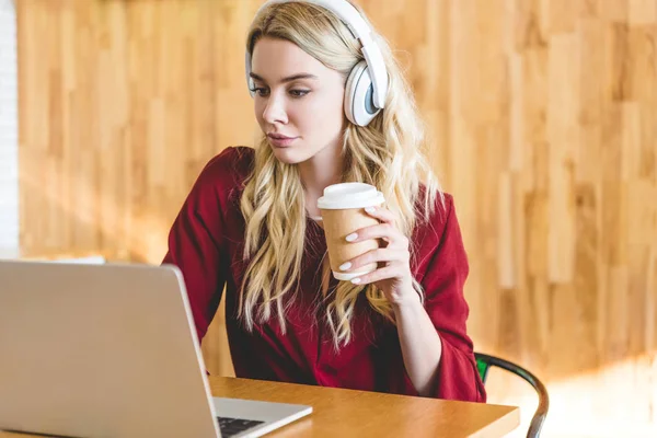 Atractiva mujer con auriculares sosteniendo la taza de papel y el uso de ordenador portátil — Stock Photo