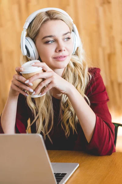 Mulher bonita com fones de ouvido segurando copo de papel e usando laptop — Fotografia de Stock