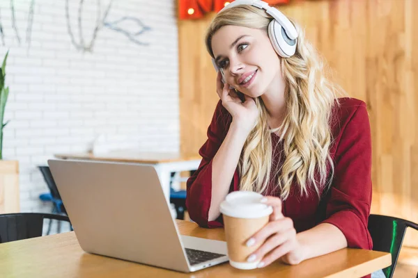 Selective focus of attractive woman listening music with headphones and holding paper cup — Stock Photo