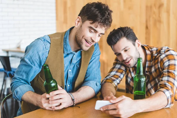 Handsome and smiling men holding glass bottles of beer and using smartphone — Stock Photo