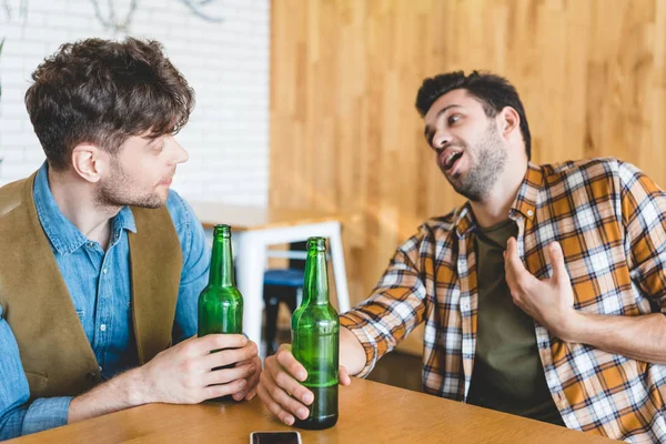 Hombres guapos sosteniendo botellas de vidrio de cerveza y hablando en la cafetería - foto de stock