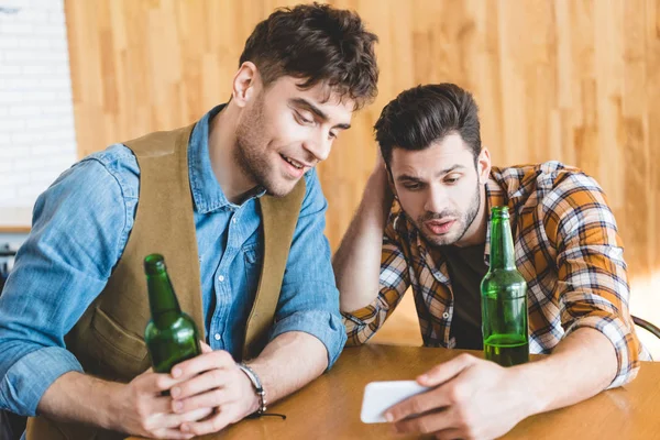 Hombres guapos sosteniendo botellas de vidrio de cerveza y el uso de smartphone - foto de stock