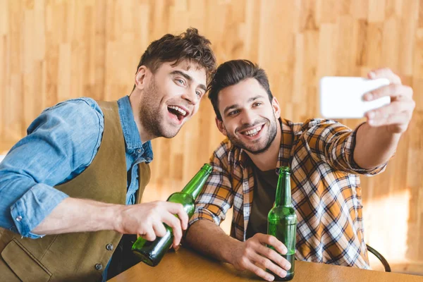Hombres guapos sosteniendo botellas de vidrio de cerveza y tomando selfie - foto de stock