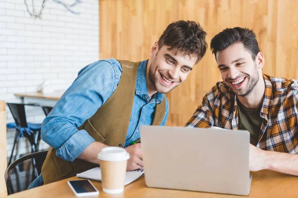 Handsome and smiling friends using laptop with paper cup on table at cafe — Stock Photo