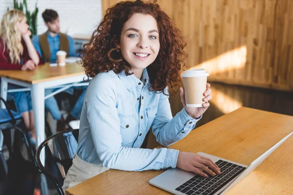 Messa a fuoco selettiva della donna attraente in possesso di tazza di carta e utilizzando il computer portatile al caffè — Foto stock