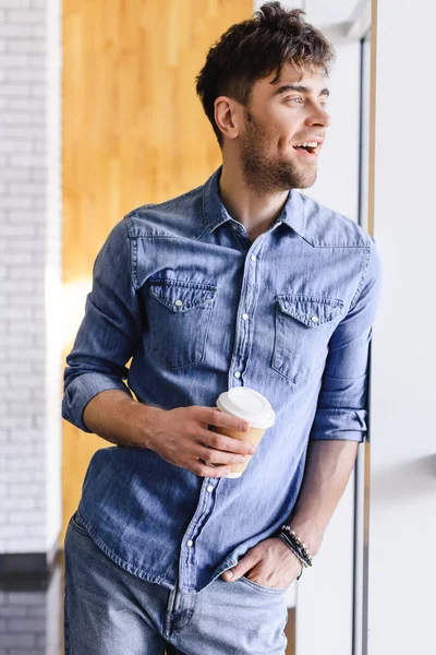 Красивый и улыбающийся мужчина стоит и держит бумажную чашку в кафе — стоковое фото