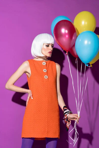 Chica pensativa en peluca blanca de pie con el brazo akimbo y la celebración de globos de aire sobre fondo púrpura - foto de stock