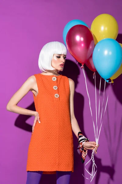 Pensive girl in white wig standing with arm akimbo and holding air balloons on purple background — Stock Photo