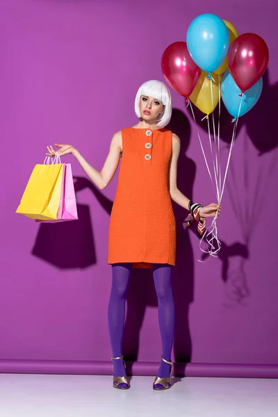 Sad girl in white wig holding shopping bags and air balloons on purple background — Stock Photo