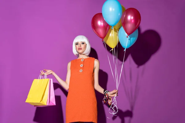 Charmantes Mädchen in weißer Perücke mit Einkaufstaschen und Luftballons auf lila Hintergrund — Stockfoto