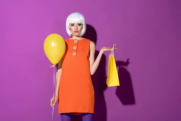 Menina elegante na peruca segurando balão de ar amarelo e saco de compras no fundo roxo — Fotografia de Stock