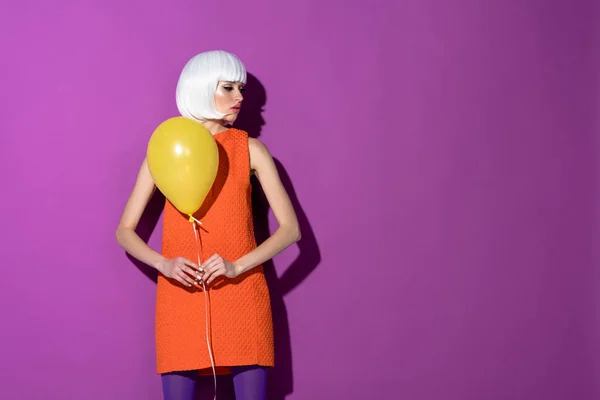 Chica en peluca blanca sosteniendo globo de aire amarillo sobre fondo púrpura - foto de stock