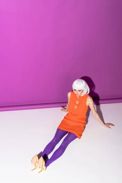 Girl in white wig and orange dress sitting on floor on purple background — Stock Photo