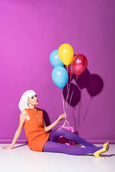 Elegante mujer joven en peluca sosteniendo globos de aire y sentado sobre fondo púrpura - foto de stock